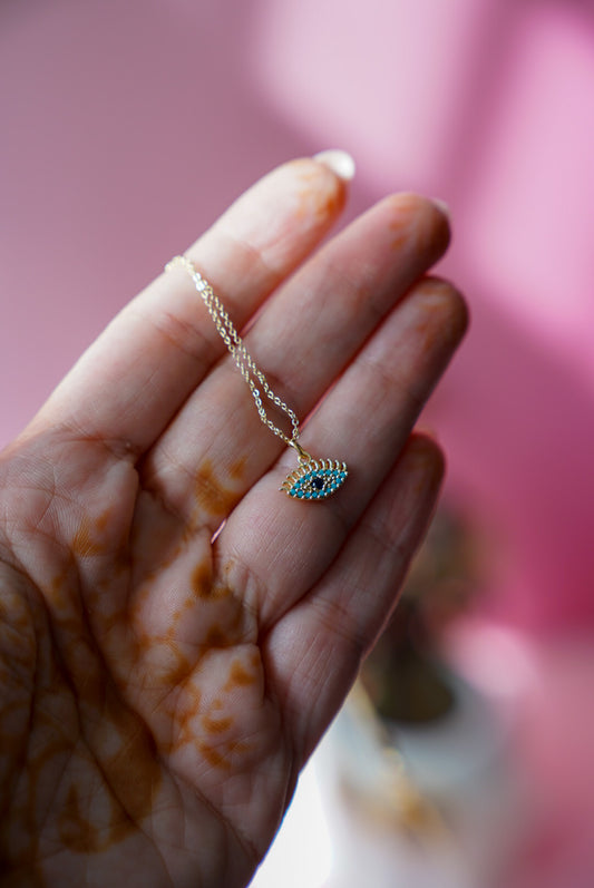 Hand holding a gold necklace with a blue eye pendant against a pink background