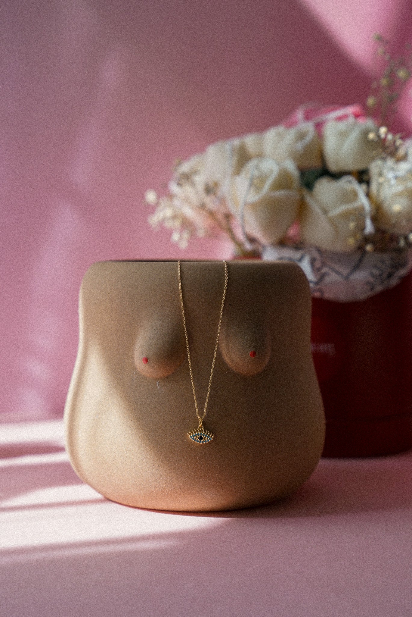 Beige female vase with a necklace draped over it on a pink surface with flowers in the background