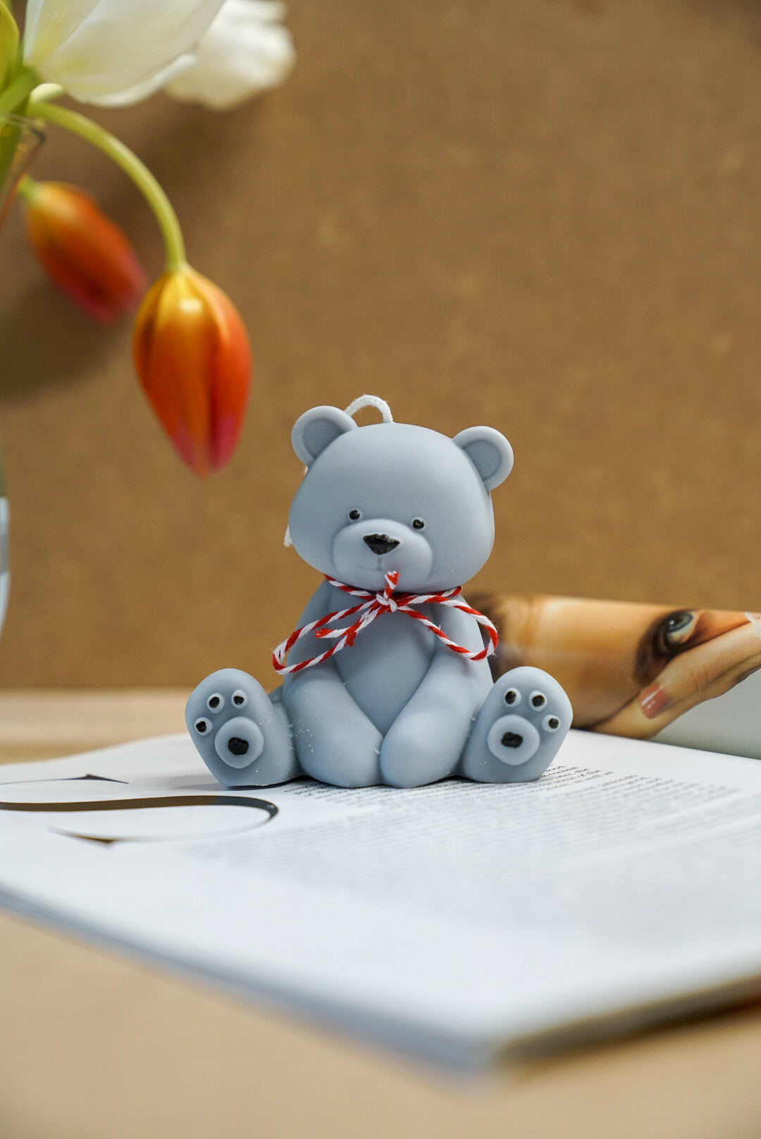 Grey Teddy Bear Candle with red and white ribbon on with tulips on the background.