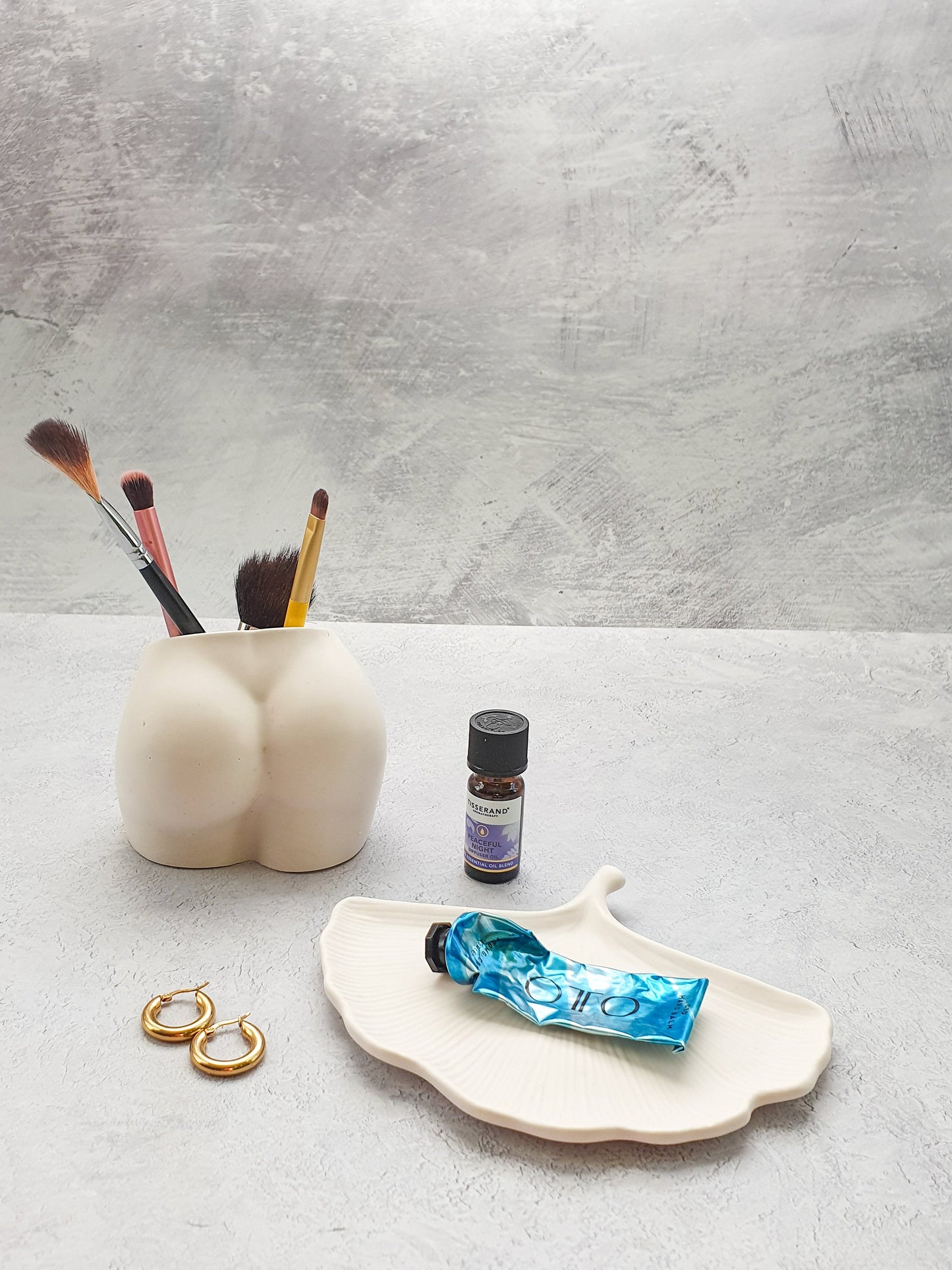 Stoneware bottom vase with some makeup brush in it with a grey background with a trinket tray and cream on it.