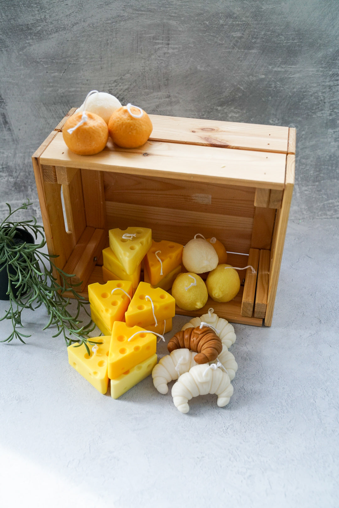 Cheese, croissant, lemon and orange shaped soy wax candles are on the wooden box with green plans on the side with a grey background.