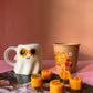Ghost-shaped mug with sunflower eyes, pumpkin-shaped candles, and a cardboard cup on a pink background.