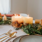 Decorative table setting with candles, greenery, and gold cutlery on a white tablecloth.